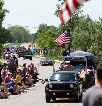 Decorated vehicles moving along the road, surrounded on either side by a crowd, during a Memorial Day parade. 